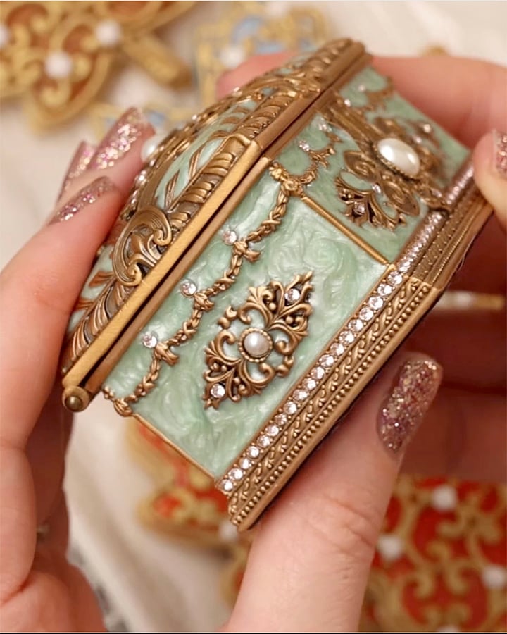 Two hands are holding a light blueish green bejeweled enameled trinket box. The hand has rose gold glitter nail polish. There are enameled fall leaf cookies out of focus in the background.