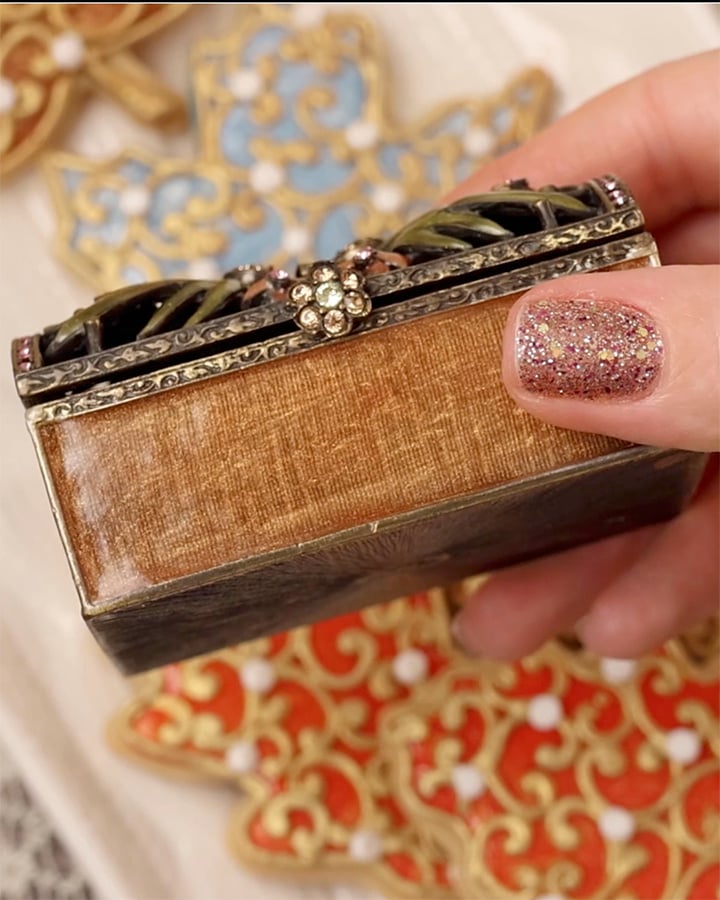 A hand is holding a brown and gold enameled trinket box. The hand has rose gold glitter nail polish. There are enameled fall leaf cookies out of focus in the background.