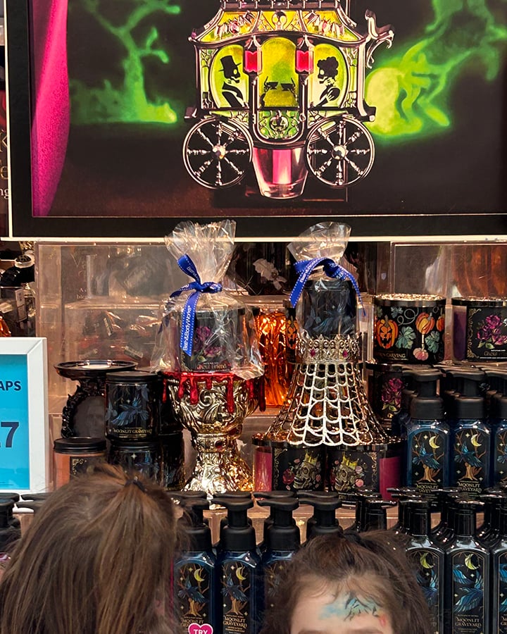 A Halloween display at bath and body works showing a golden carriage with two skeleton silhouettes inside, rows of black soap bottles, and two golden candle holders that look like a goblet and a spider web.
