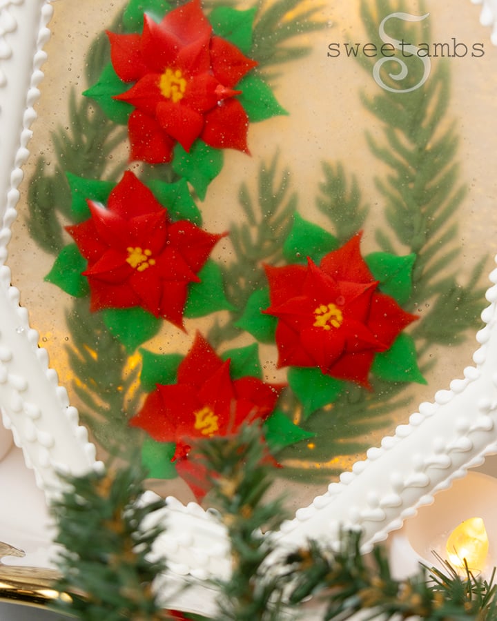 A close up of the edible holiday centerpiece with royal icing poinsettias and royal icing evergreen branches encased in isomalt inside of a white octagon shaped cookie frame covered in royal icing. The centerpiece is in front of a golden bokeh lights background and there are artificial evergreen boughs on the table in front of the centerpiece.