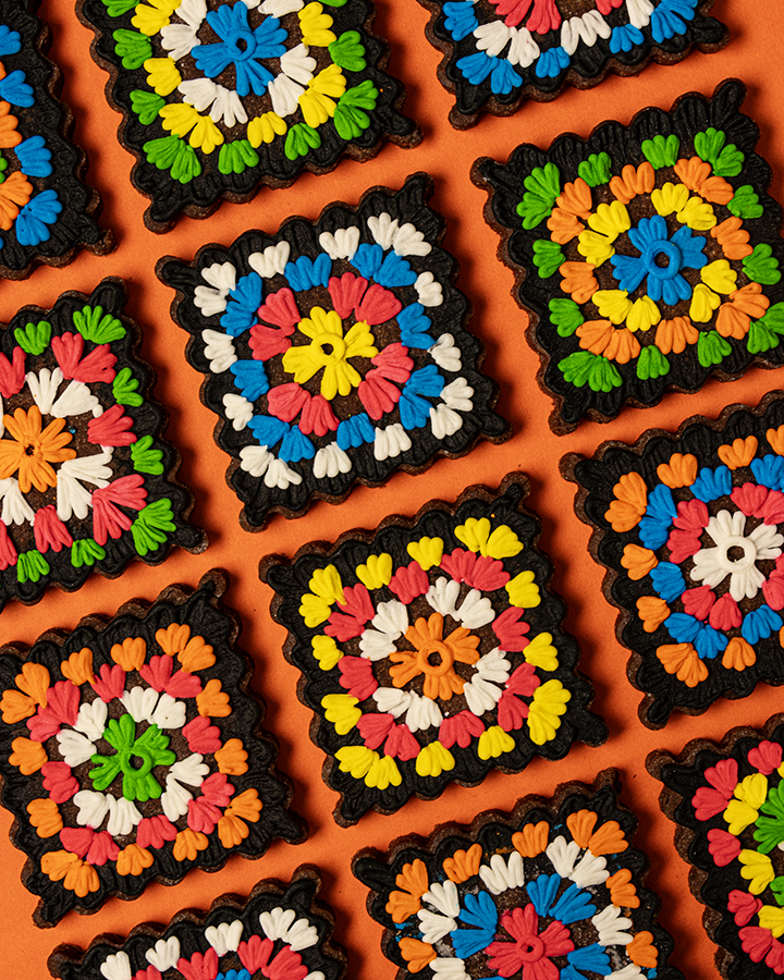 A group of square fluted edged cookies decorated with black and brightly colored icing to look like crochet granny squares. The cookies are on an orange background.