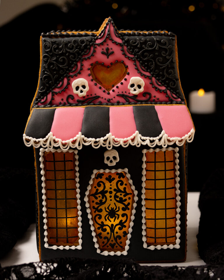 A gingerbread house decorated in a Halloween style with pink and black icing, a scalloped awning, royal icing skulls, sugar glass windows, and piped filigree decorations.