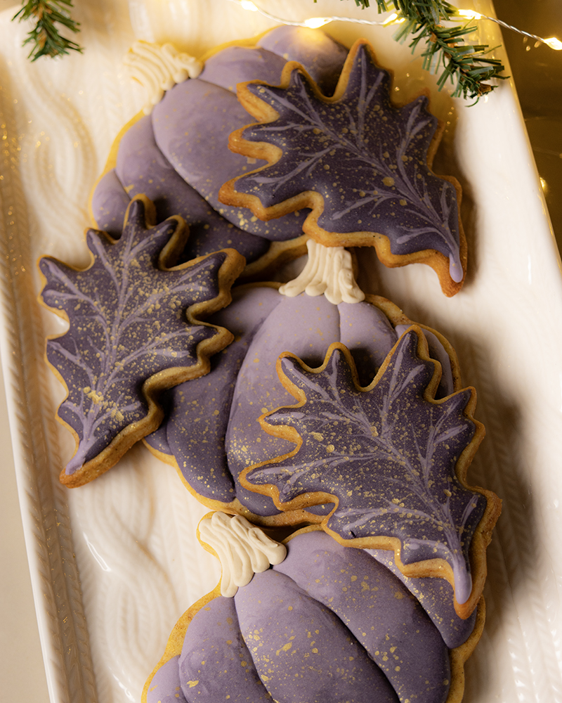 Elaborate plum and gold decorated sugar cookies, shaped as pumpkins and fall leaves, on a white platter with a cable knit design.