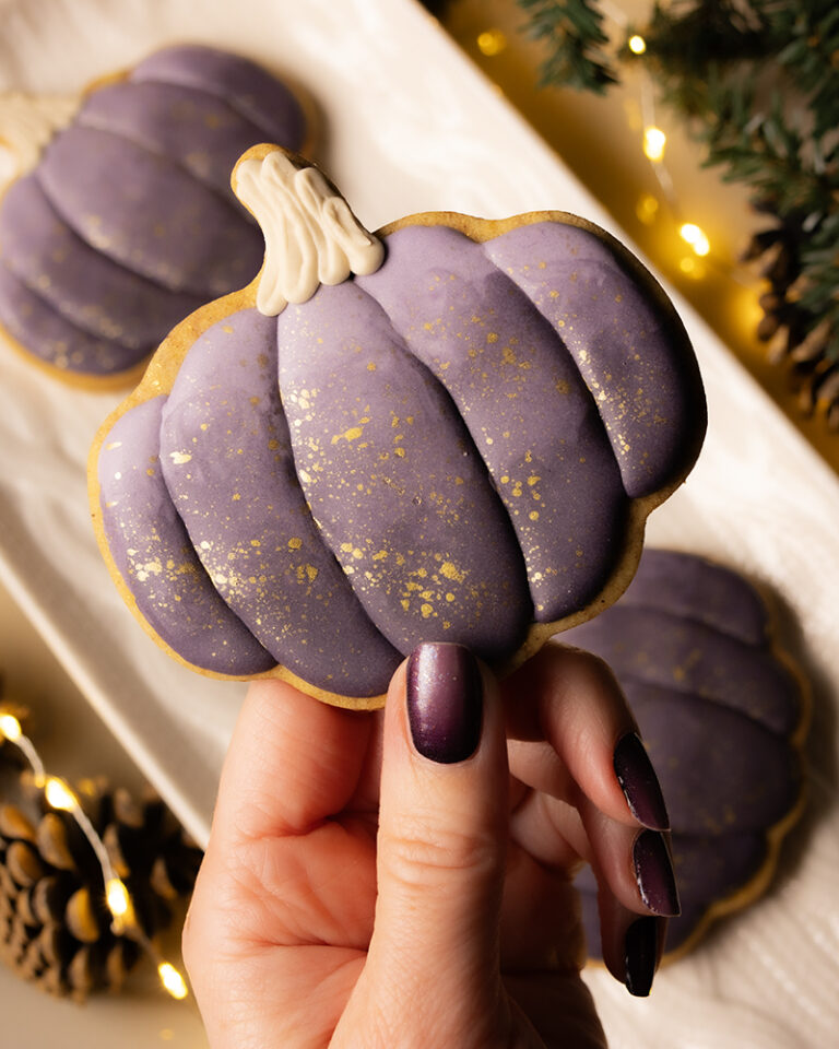 A pumpkin shaped cookie decorated with a deep shade of plum icing. There are gold speckles on the icing. The cookie is being held by a hand with nails that are painted with a similar shade. There is a platter of more pumpkin cookies in the background surrounded by fairy lights and evergreen branches.
