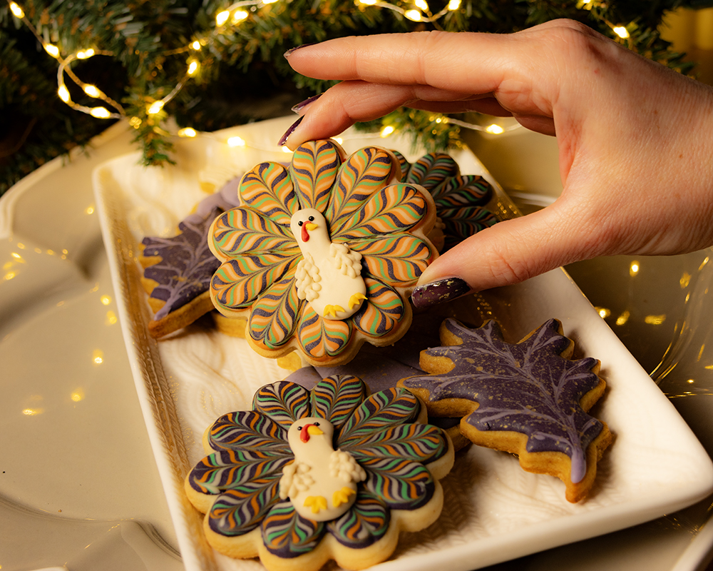 Royal icing Thanksgiving cookies decorated as a colorful turkey with vibrant tail feathers, purple pumpkins, and dark purple maple leaves with gold speckles, displayed on a platter. A hand with dark purple nail polish is holding the turkey cookie.