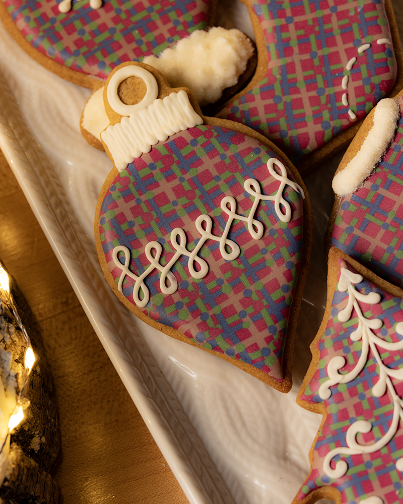 A close-up view of a Christmas ornament-shaped gingerbread cookie resting on a white serving platter. The cookie is decorated with a vibrant, multicolored wet-on-wet royal icing plaid pattern, and a horizontal line of white swirled piping runs across the center. The top of the ornament features a ribbed white royal icing cap. Other plaid cookies and string lights are visible around it, emphasizing a festive setting.