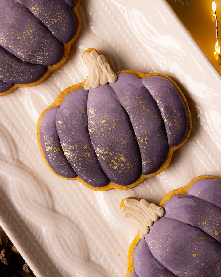 pumpkin shaped cookies decorated with a deep shade of plum icing. There are gold speckles on the icing. The cookies are on a white platter with a cable knit design. There are fairy lights around the platter.
