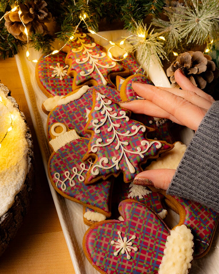 A close-up shot of a white platter filled with elaborately decorated Christmas cookies, illuminated by warm string lights. The cookies are cut into festive shapes like gingerbread men, Christmas trees, hearts, and mittens. Many are decorated with a vibrant, multicolored, wet-on-wet royal icing pattern resembling a festive plaid or tartan. A hand wearing a gray sweater reaches for a Christmas tree-shaped cookie, which is decorated with delicate white royal icing details that look like snowy branches. Pine sprigs and pinecones decorate the background.
