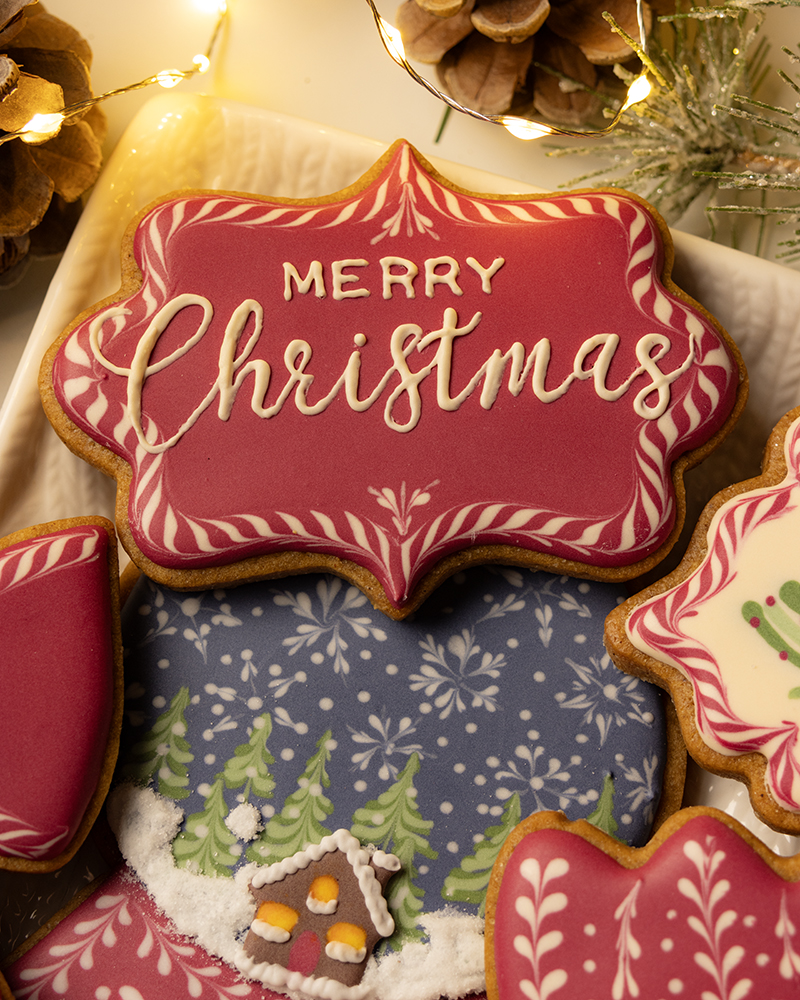 A close-up shot of several Christmas cookies on a white platter, with a central red, plaque-shaped cookie featuring the white, hand-piped text "Merry Christmas" and wet-on-wet scrollwork. The background includes pinecones, evergreen sprigs, and warm fairy lights.