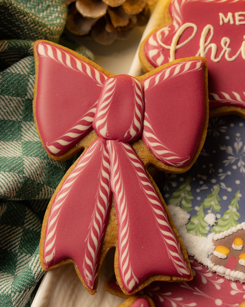 A gingerbread cookie shaped like a large red bow, decorated with red and white candy cane-striped wet-on-wet designs on the tails and center knot. It sits on a white platter beside a dark green and white checkered napkin, with pinecones and other cookies visible in the background.