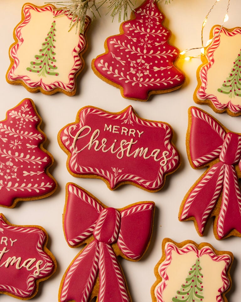 A top-down shot of a collection of intricately decorated Christmas sugar cookies, featuring red and white royal icing in various shapes, with a sprig of frosted evergreen foliage and a string of warm fairy lights visible at the top.