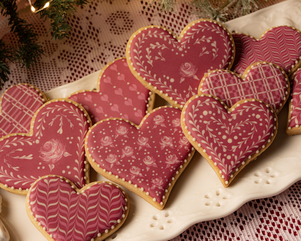 Decorated pink heart-shaped sugar cookies featuring floral and filigree white icing patterns, arranged on a white platter with a lace tablecloth background