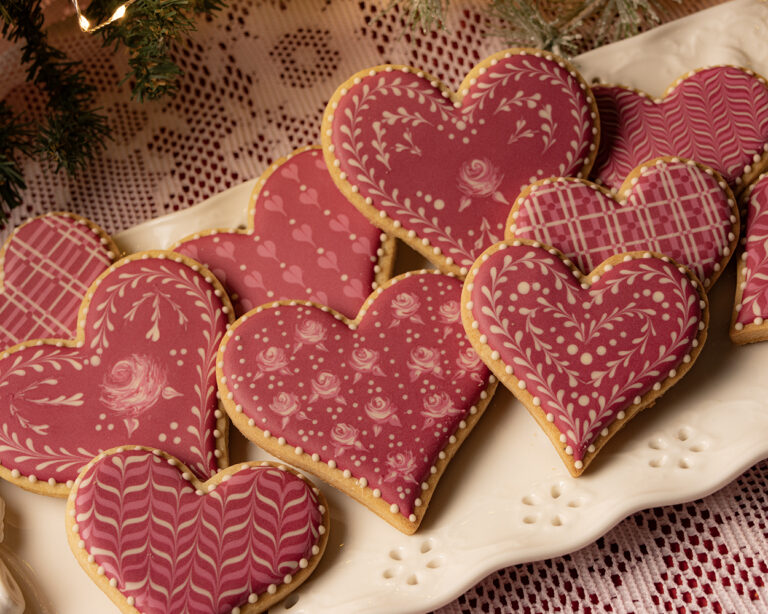 Decorated pink heart-shaped sugar cookies featuring floral and filigree white icing patterns, arranged on a white platter with a lace tablecloth background
