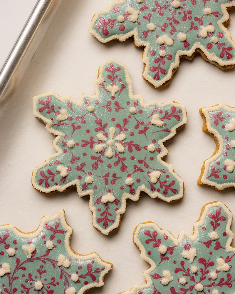 A close up of Snowflake-shaped sugar cookies decorated with pale blue royal icing, red snowflake patterns, and small white icing dots, arranged in a neat pattern on a light background.