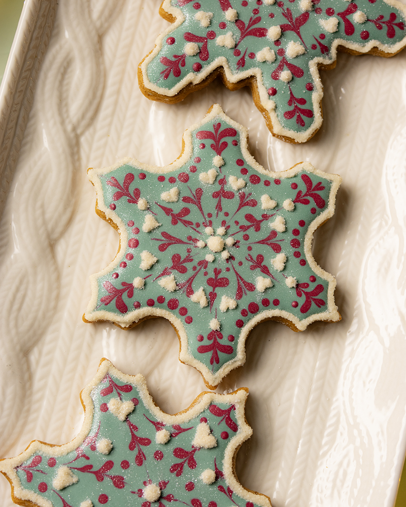 A close up of snowflake-shaped sugar cookies decorated with pastel blue royal icing and red filigree details, arranged on a white platter.