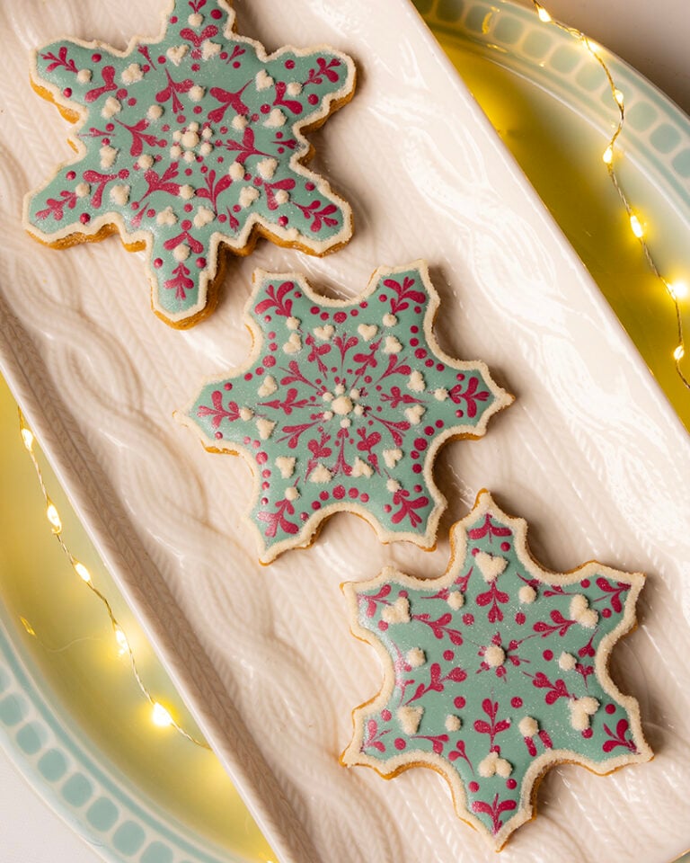 Three snowflake-shaped sugar cookies decorated with pastel blue royal icing and red filigree details, arranged on a white platter with soft glowing string lights.