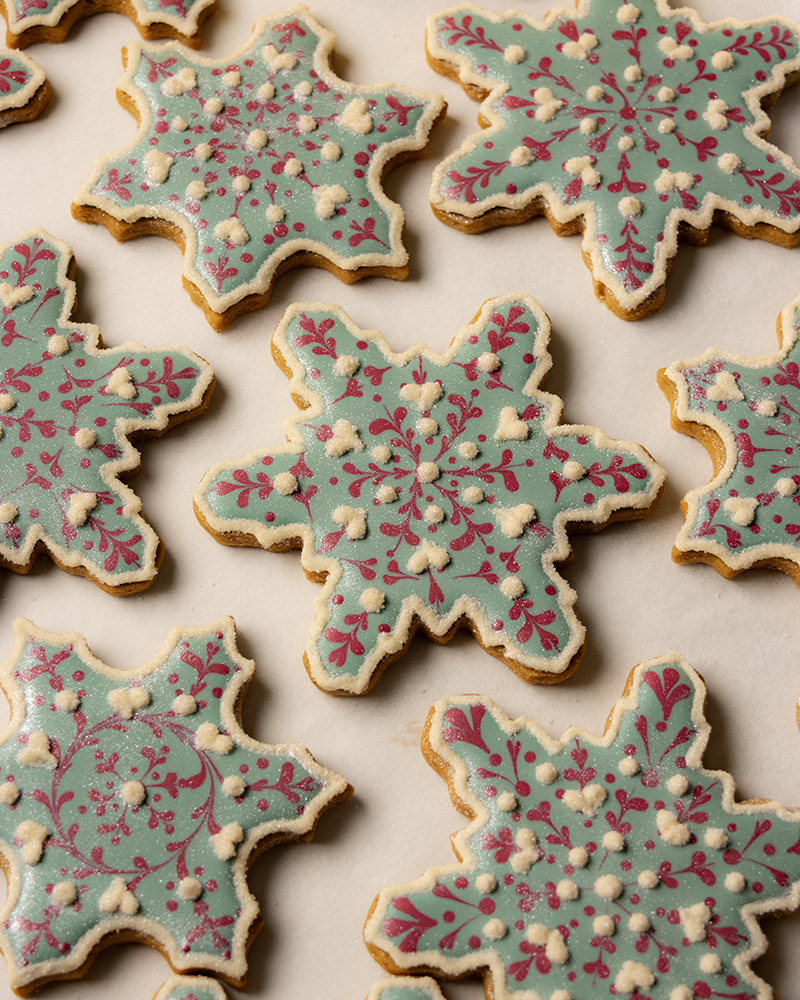 Snowflake-shaped sugar cookies decorated with pale blue royal icing, red snowflake patterns, and small white icing dots, arranged in a neat pattern on a light background.