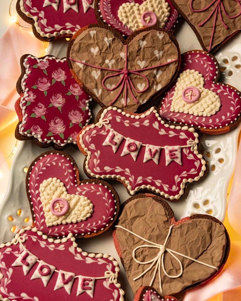 Pink, brown, and cream heart-shaped and plaque-shaped Valentine’s cookies with detailed icing patterns, bows, and “LOVE” text, displayed together on a white platter.