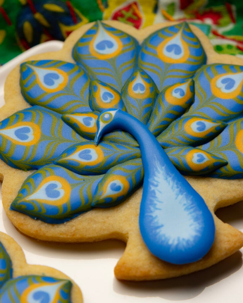 A close up of a peacock-shaped cookie with a blue body and layered blue, green, and yellow royal icing feathers arranged on a white plate.