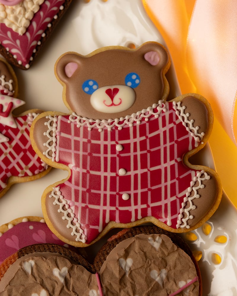 Teddy bear–shaped Valentine’s Day cookie decorated with brown icing and blue eyes, wearing a red plaid sweater with white lace details, displayed on a white plate with other heart-shaped cookies.