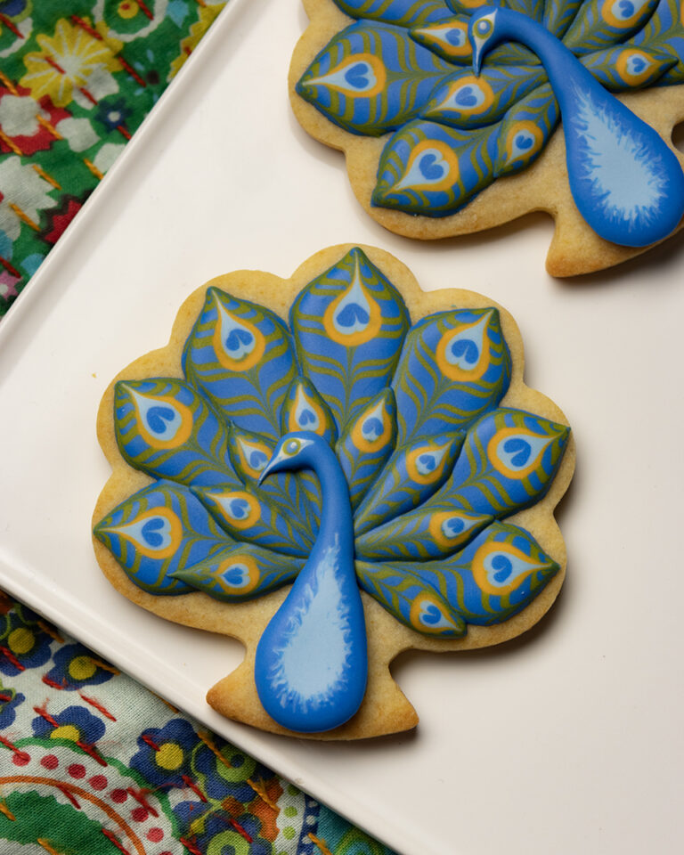 Peacock-shaped cookie with a blue body and layered blue, green, and yellow royal icing feathers arranged on a white plate