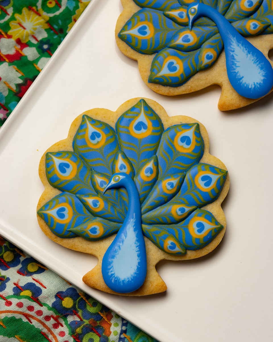 Peacock-shaped cookie with a blue body and layered blue, green, and yellow royal icing feathers arranged on a white plate