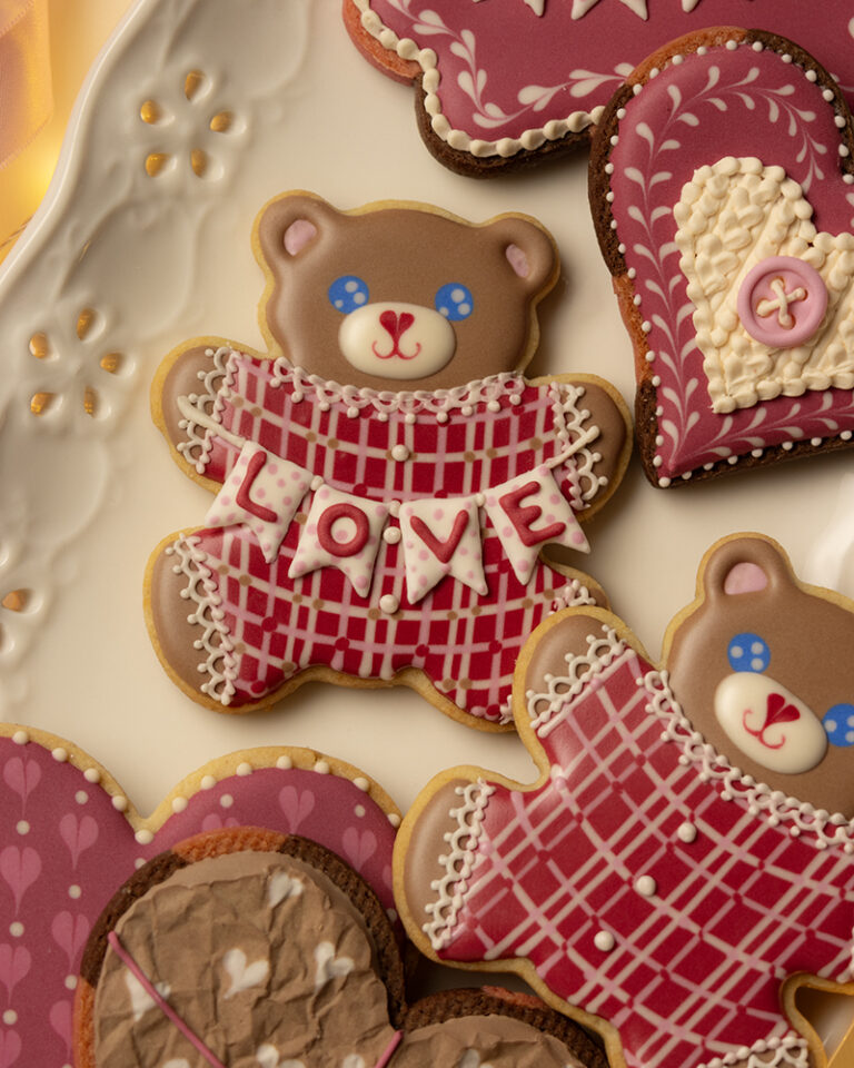 Teddy bear–shaped sugar cookie with brown icing, blue eyes, and a red plaid sweater, holding a white banner that spells “LOVE,” arranged on a plate with pink and heart-shaped decorated cookies.