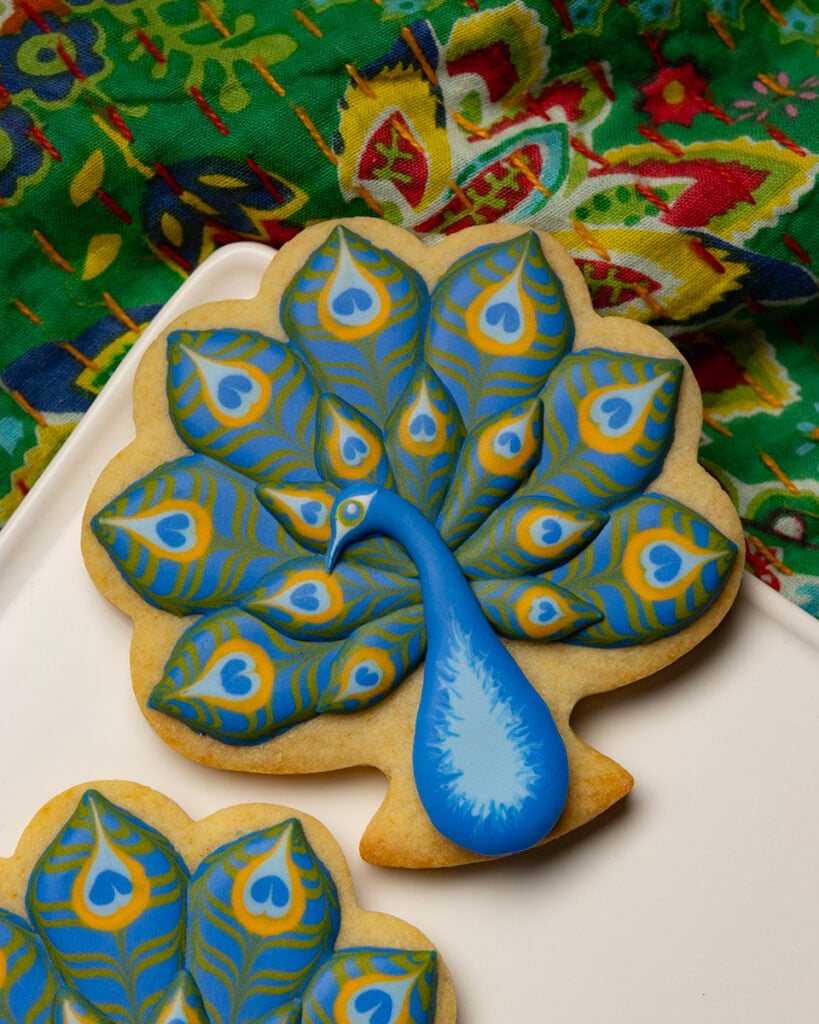 Peacock-shaped cookie with a blue body and layered blue, green, and yellow royal icing feathers arranged on a white plate over a colorful quilt.