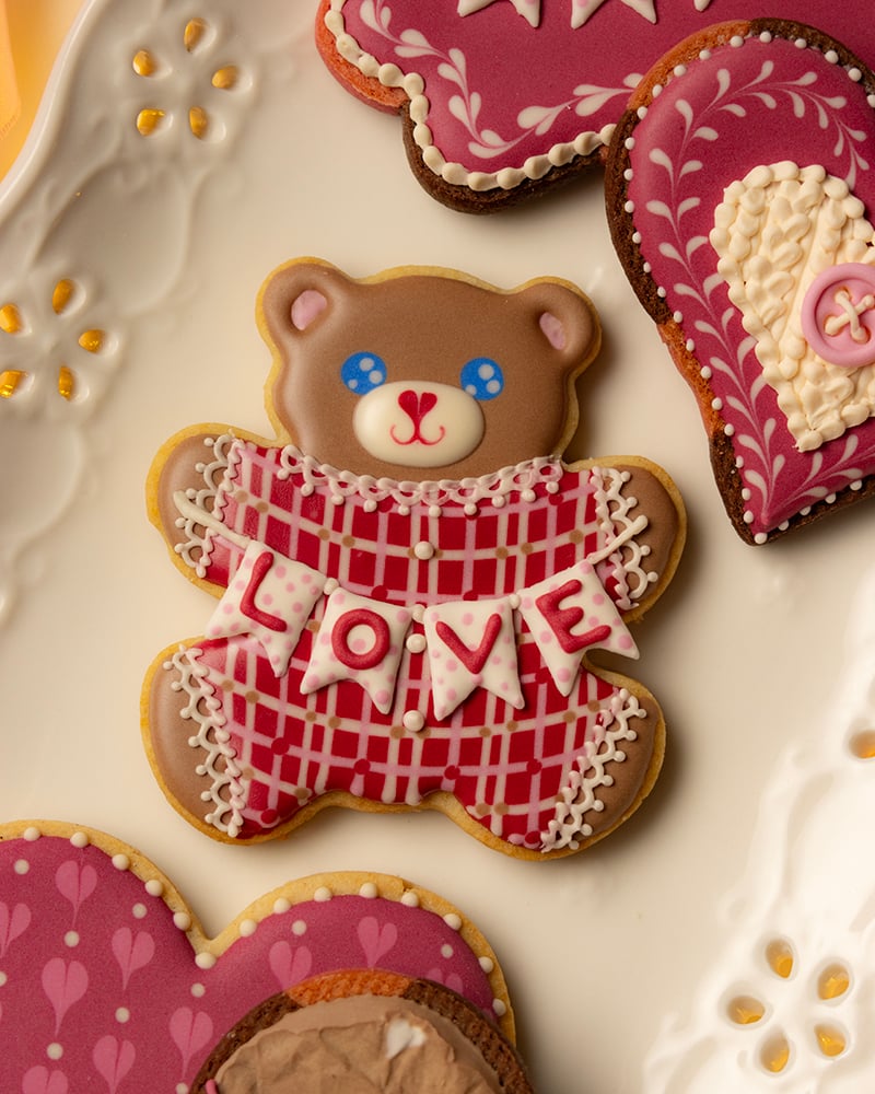 Teddy bear–shaped sugar cookie with brown icing, blue eyes, and a red plaid sweater, holding a white banner that spells “LOVE,” arranged on a plate with pink and heart-shaped decorated cookies.