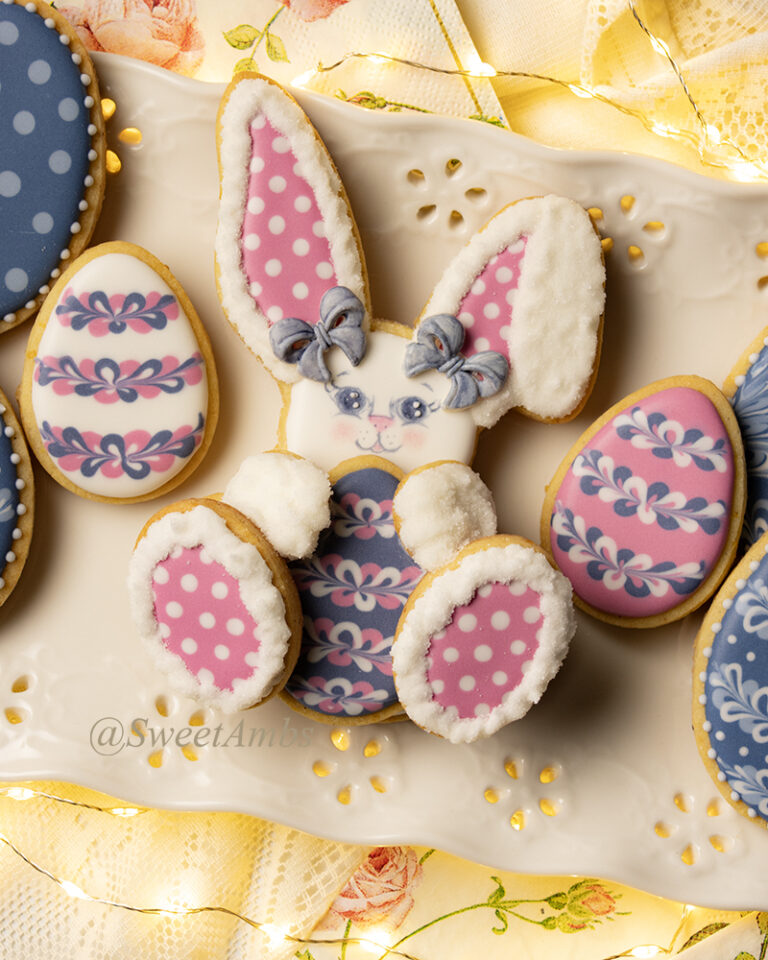 An easter bunny cookie with pink polka dotted ears and feet holds a decorated easter egg cookie. The cookie is surrounded by more decorated easter cookies. All of the cookies are on a platter with an eyelet lace design. The platter is on a lacy floral background with fairy lights.
