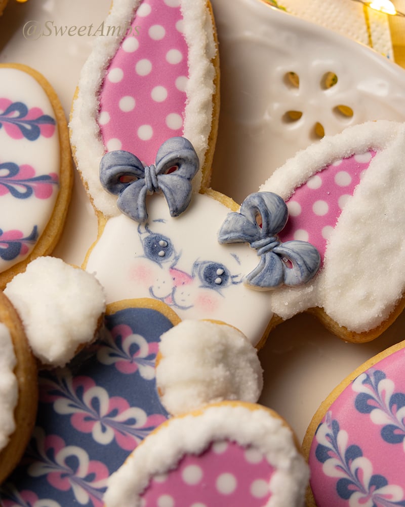 A close up of an easter bunny cookie with pink polka dotted ears and feet holds a decorated easter egg cookie. All of the cookies are on a platter with an eyelet lace design