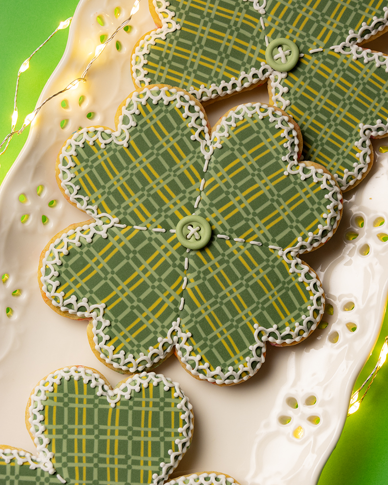 4 leaf clover shaped cookies decorated with royal icing to look like a plaid stitched quilt with a button in the middle. The cookies are on a platter with an eyelet lace design on the edge. The plater is on a green background and surrounded by fairy lights.