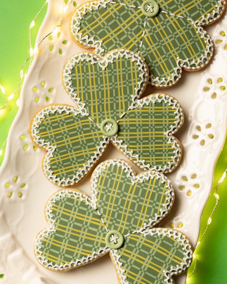 Shamrock shaped cookies decorated with royal icing to look like a plaid stitched quilt with a button in the middle. The cookies are on a platter with an eyelet lace design on the edge. The plater is on a green background and surrounded by fairy lights.