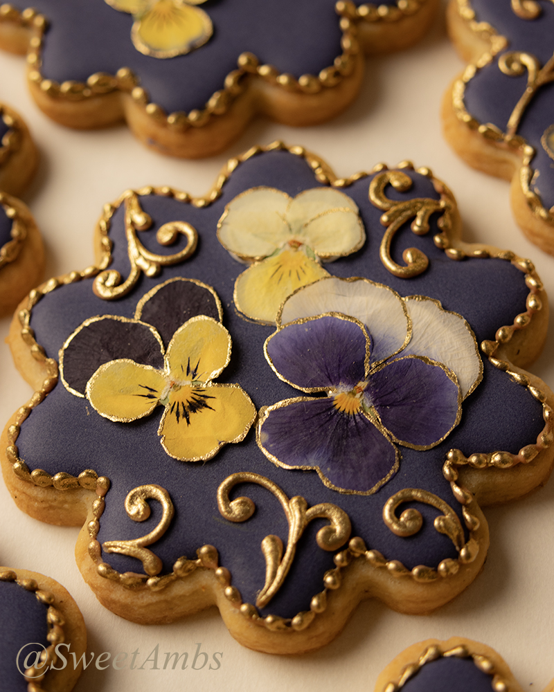 Round scalloped edge cookies decorated with a deep purple icing base layer, dried pressed violas, and gold filigree.