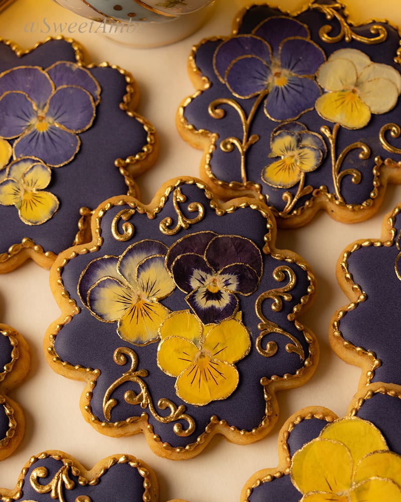 Round scalloped edge cookies decorated with a deep purple icing base layer, dried pressed violas, and gold filigree.