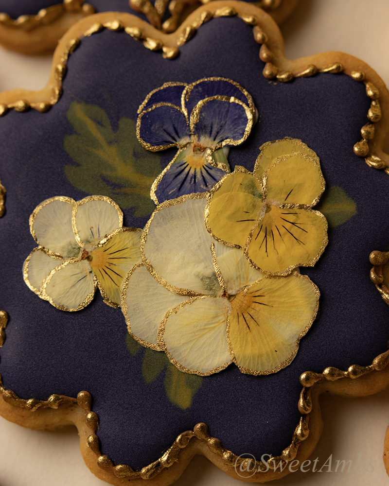 A close up of the round scalloped edge cookies decorated with a deep purple icing base layer, dried pressed violas, and a gold border.