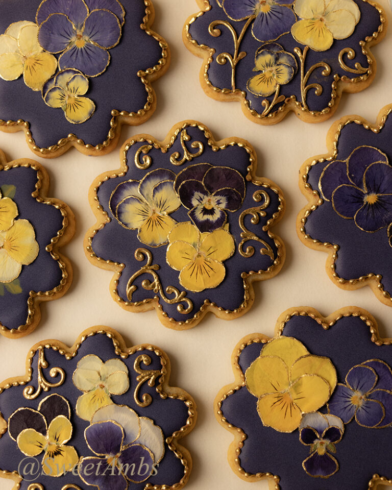 Round scalloped edge cookies decorated with a deep purple icing base layer, dried pressed violas, and gold filigree.