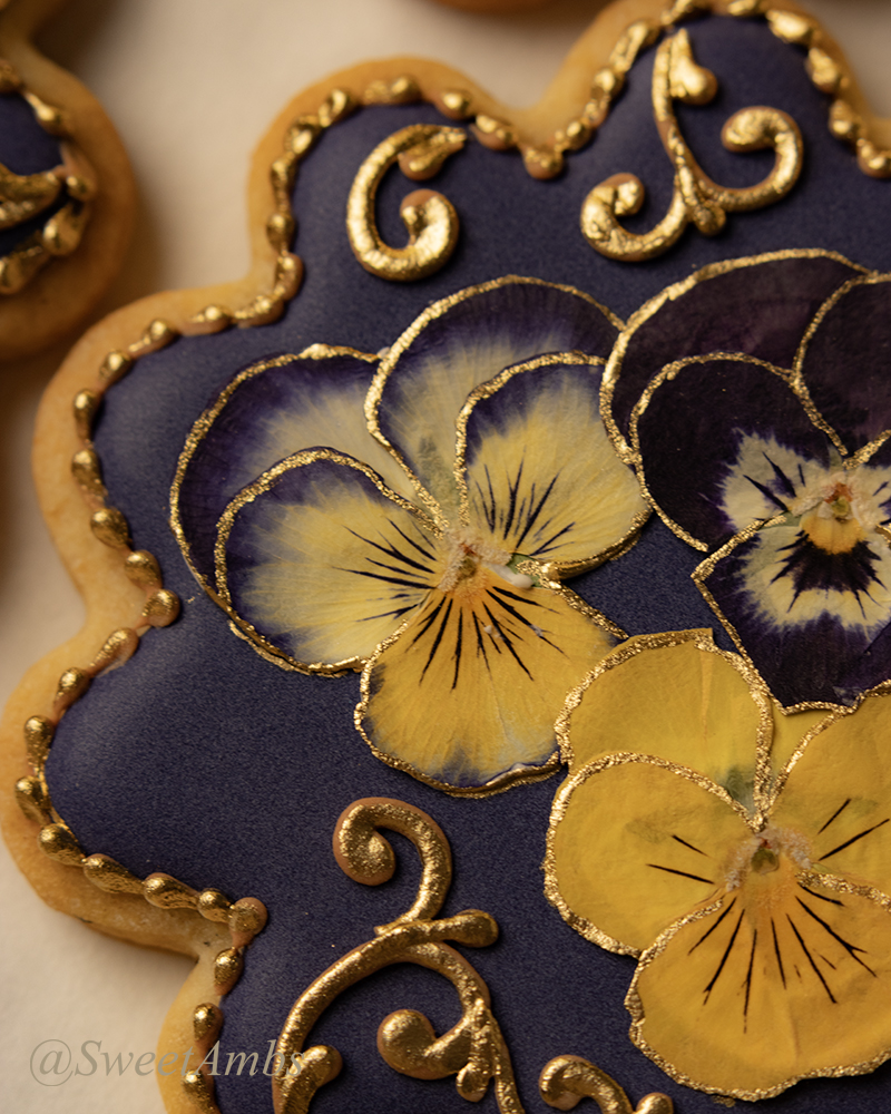 A close up of the round scalloped edge cookies decorated with a deep purple icing base layer, dried pressed violas, and gold filigree.