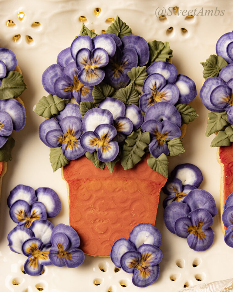A cookie decorated to look like a bunch of violas in a terra cotta pot. There are a few royal icing violas surrounding the cookie. The cookie is on a platter with an eyelet lace design on the edge.