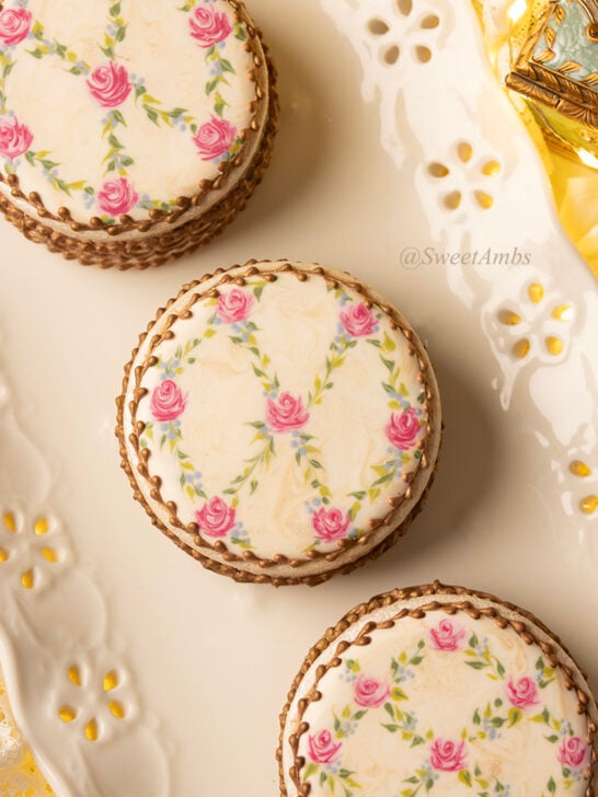 Cookies decorated to resemble vintage floral enameled makeup compacts. There are 3 cookie boxes on a plate and one of the lids is askew, revealing bronzed chocolate chips inside. The platter has an eyelet lace edge and there are fairy lights in the background.