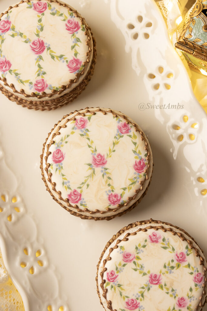 Cookies decorated to resemble vintage floral enameled makeup compacts. There are 3 cookie boxes on a plate and one of the lids is askew, revealing bronzed chocolate chips inside. The platter has an eyelet lace edge and there are fairy lights in the background.