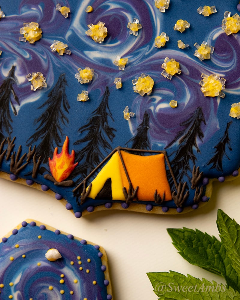 A close up of the summer camping cookie showing an orange and yellow tent and a small campfire among the pine trees with a Starry Night inspired swirled sky.