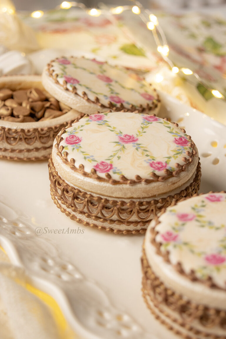 Cookies decorated to resemble vintage floral enameled makeup compacts. There are 3 cookie boxes on a plate and one of the lids is askew, revealing bronzed chocolate chips inside. The platter has an eyelet lace edge and there are fairy lights in the background.
