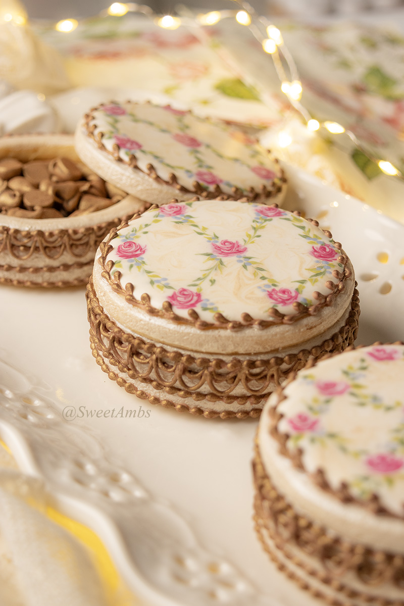 Cookies decorated to resemble vintage floral enameled makeup compacts. There are 3 cookie boxes on a plate and one of the lids is askew, revealing bronzed chocolate chips inside. The platter has an eyelet lace edge and there are fairy lights in the background.