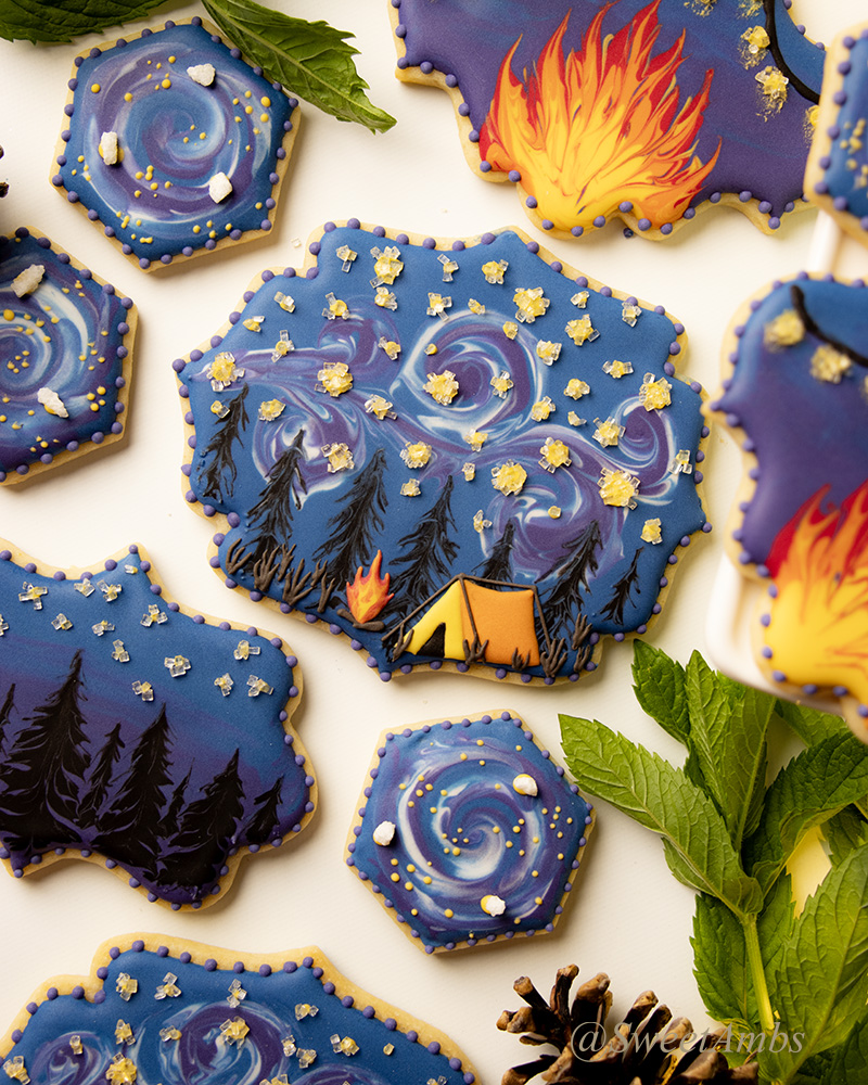 A set of decorated cookies in plaque shapes and small hexagons decorated with deep blue, purple, and black icing depicting a camping scene with stars, campfires, and pine trees. There is a sprig of mint and a pine cone in the corner.