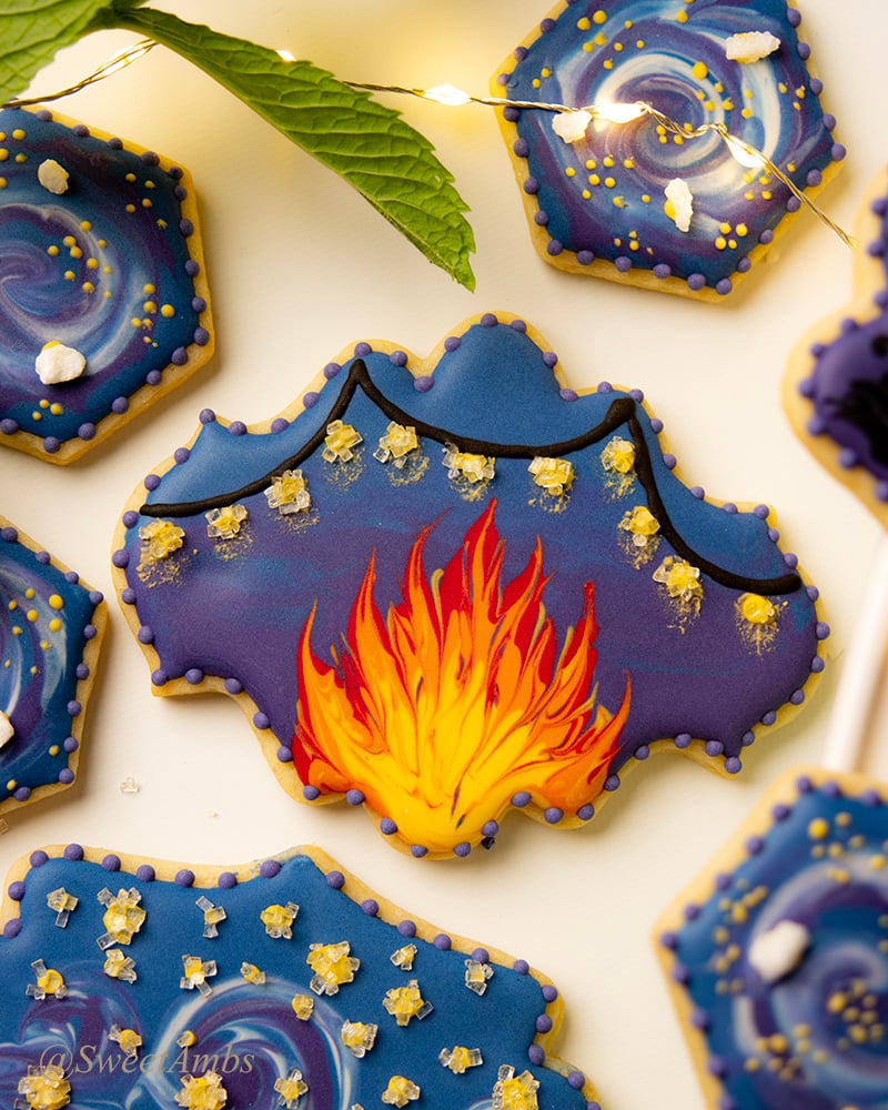 A plaque shaped cookie decorated with a dark blue and purple background with a campfire and string lights. The cookie is surrounded by other camping themed decorated cookies, fairy lights, and a sprig of mint.