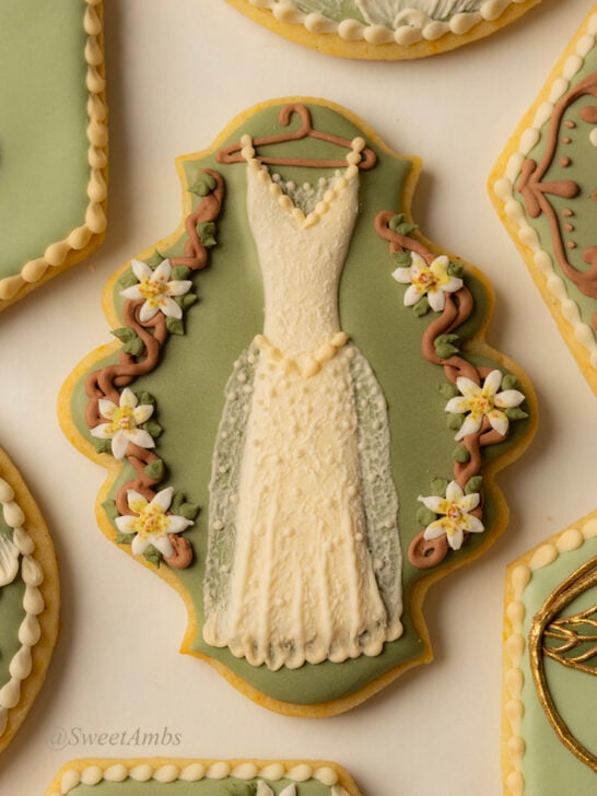 A plaque shaped cookie decorated with green icing and a lacy wedding dress on a hanger surrounded by vines and white flowers.