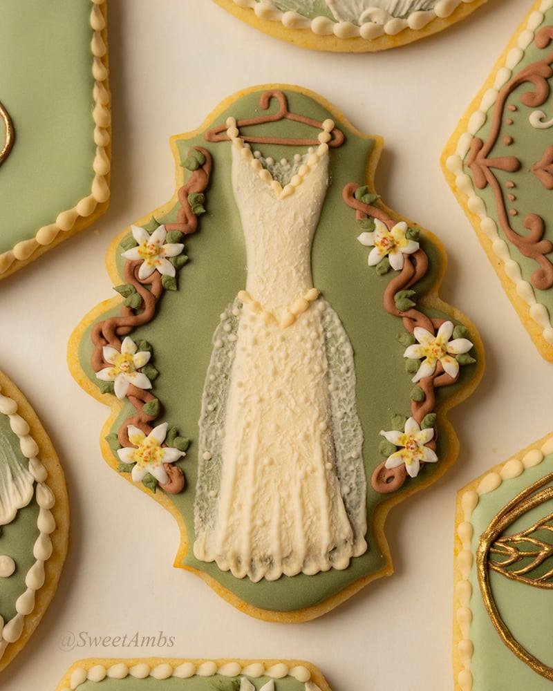 A plaque shaped cookie decorated with green icing and a lacy wedding dress on a hanger surrounded by vines and white flowers.