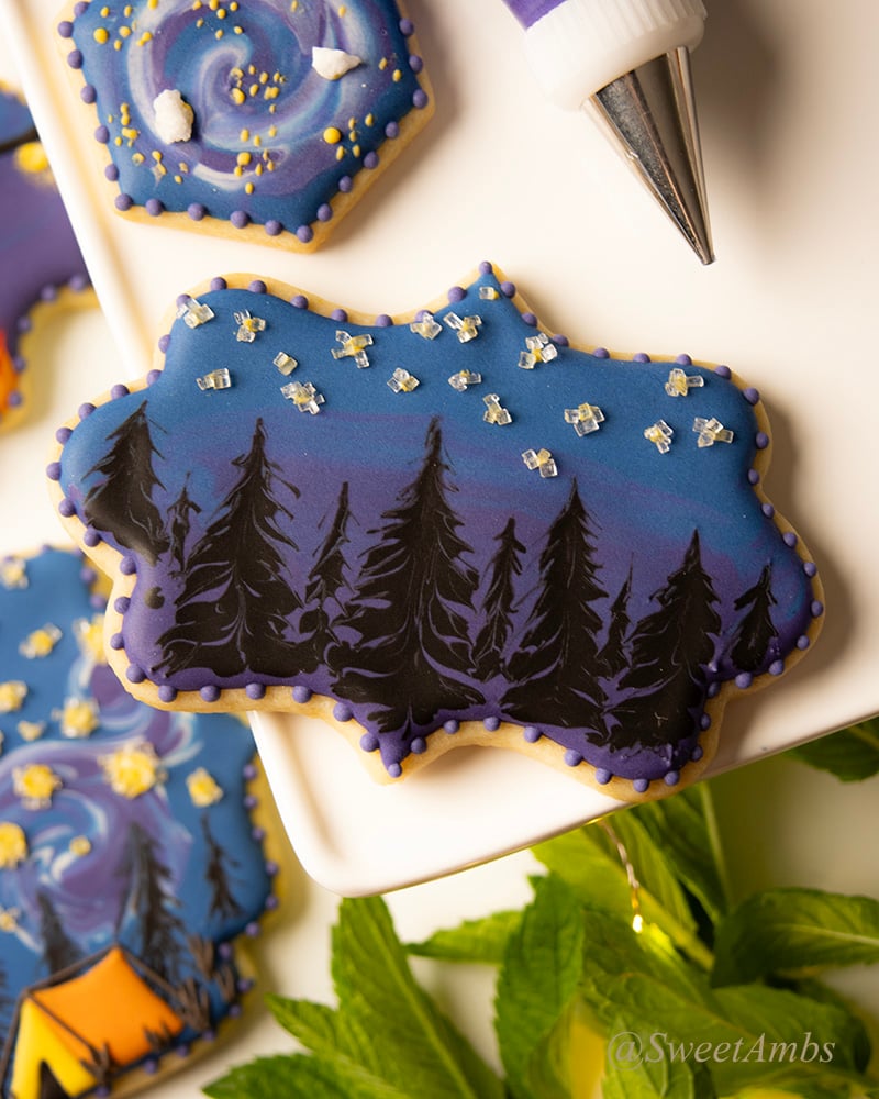 A plaque shaped cookie decorated with dark blue and purple icing with a black silhouette of a pine tree forest. There are sparkling sugar stars in the sky above the trees. The cookie is surrounded by other decorated cookies, a piping bag, and a sprig of mint.