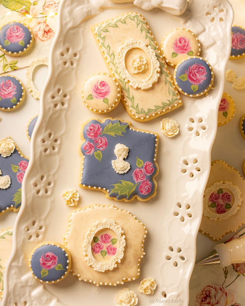 3 plaque shaped cookies decorated with florals, frames, and a cameo. The cookies are on a platter with an eyelet lace design on the edge. There are more floral cookies beneath the platter.
